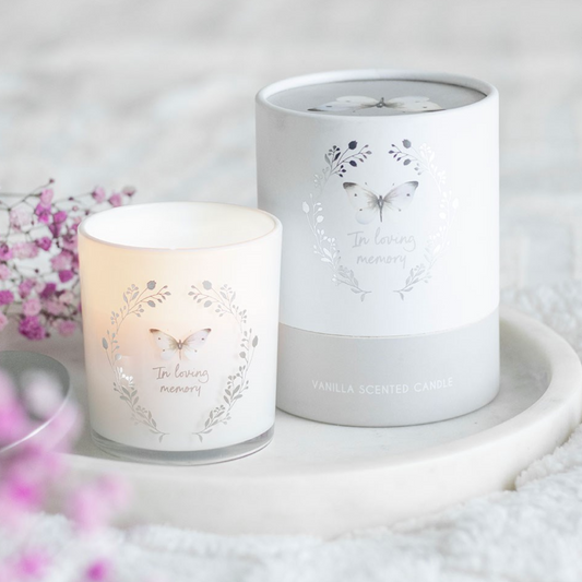 A white vanilla scented memorial candle with an “In Loving Memory” butterfly design beside its matching grey gift box on a soft neutral background.