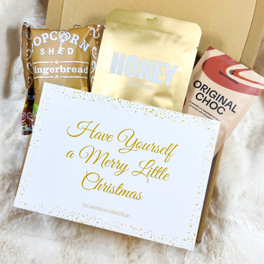 Holiday Hug Christmas Care Box displayed on a soft, fluffy background with gingerbread popcorn, honey face mask, hot chocolate and a festive handwritten Christmas notecard.