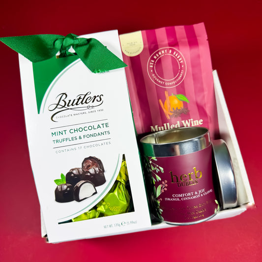 Christmas gift box featuring Butlers mint chocolate truffles, mulled wine gummies, and a Herb Dublin Comfort & Joy candle arranged on a festive red background