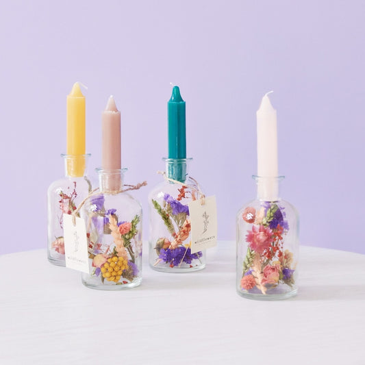 Four glass jars with dried flowers and lit candles against a light purple background